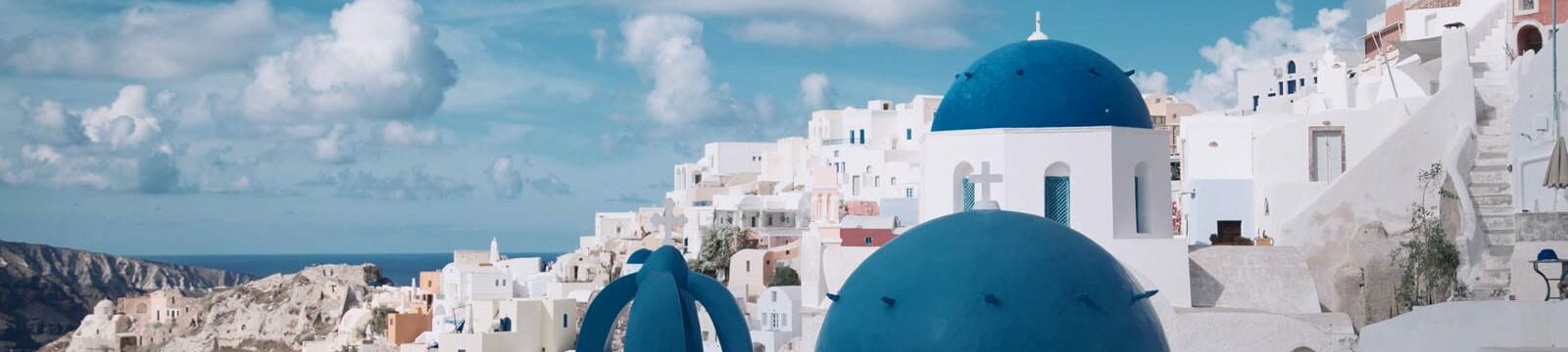 pexels-jimmy-teoh-1010657 A picturesque view of Oia, Santorini featuring iconic white-washed buildings and distinctive blue-domed churches against a backdrop of blue sky and sea.