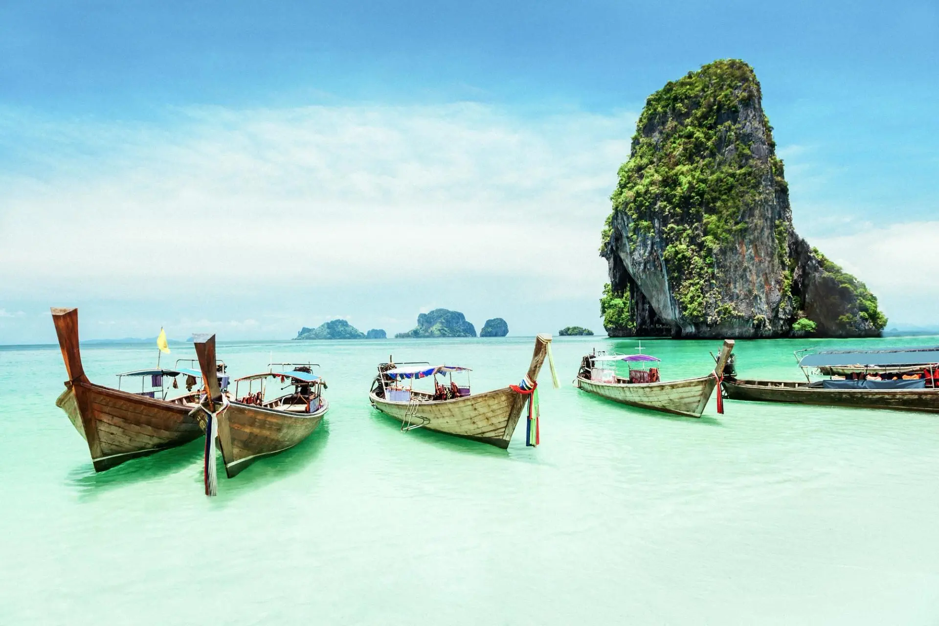 0579751453243971b2759c777f51c9f93c9a30dd-5616×3744-1 Traditional longtail boats anchored in crystal-clear turquoise water with dramatic limestone karst cliffs covered in lush vegetation rising in the background.
