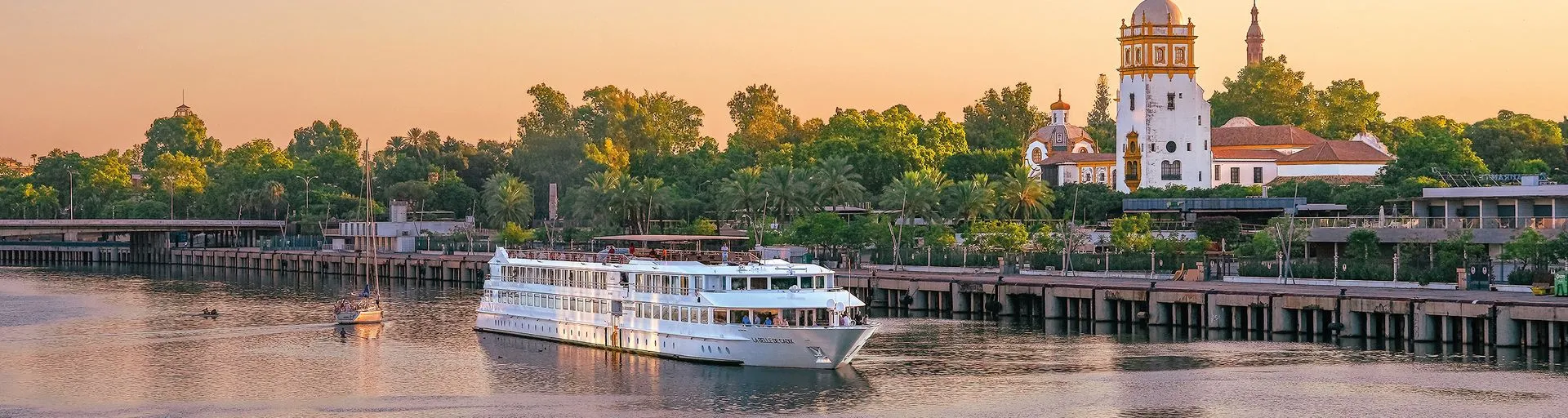1920×512-2 A multi-deck white riverboat moored at a wooden pier along a wide river during golden hour, with historic architecture visible on the bank.