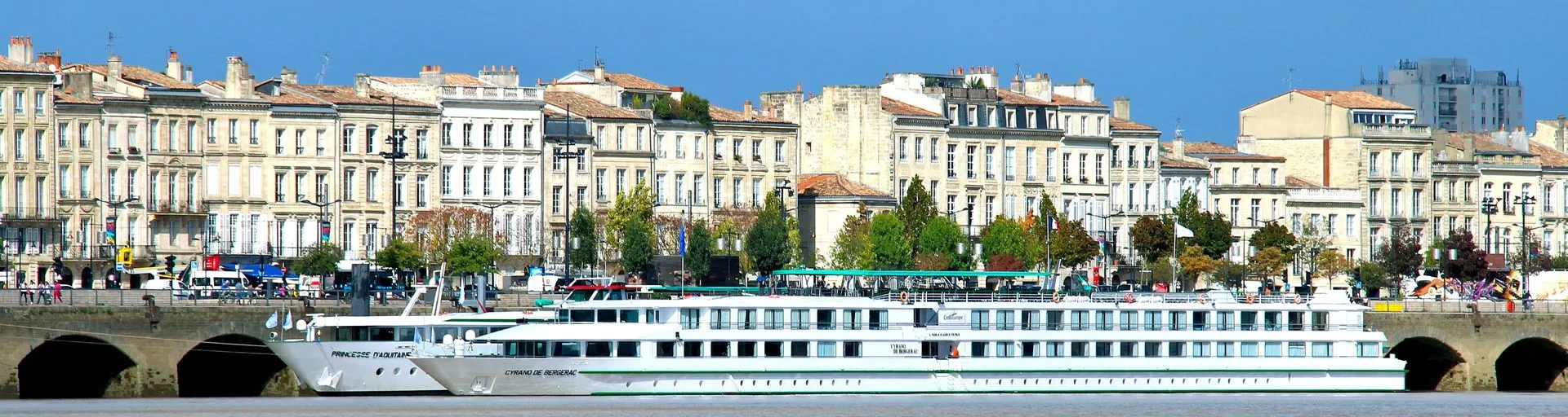 1920×512 A modern river cruise ship moored along the Garonne River in Bordeaux, France, with classic 19th-century Haussmann-style buildings lining the waterfront.