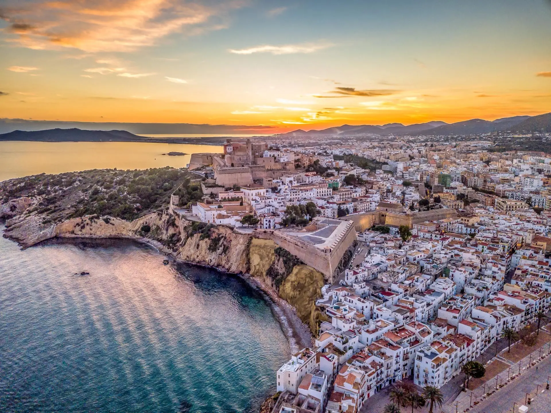 31947b0006326a3aee3322b7ea4dfcb7c2bc800a-3992×2992-1 Aerial view of Denia Castle perched on a rocky peninsula overlooking the Mediterranean Sea at sunset, with white-washed buildings and mountains in the distance.