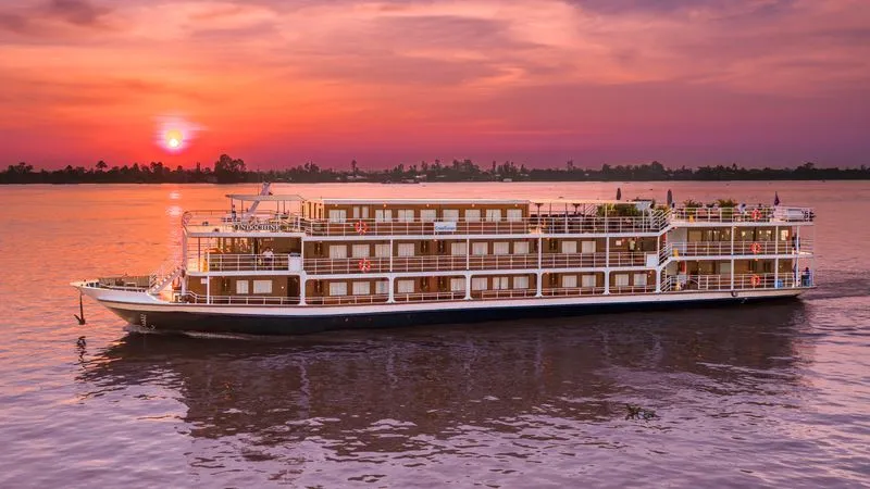 800×450-4 A multi-deck river cruise ship anchored on a calm river at golden hour, with a stunning sunset painting the sky in shades of orange, pink, and purple.
