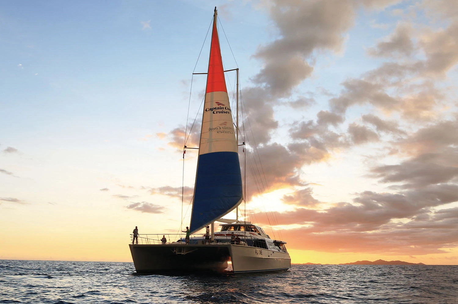 Captain Cook’s Catamaran Sunset Cruise A Captain Cook Cruises catamaran with distinctive red, tan, and blue sails glides across calm ocean
