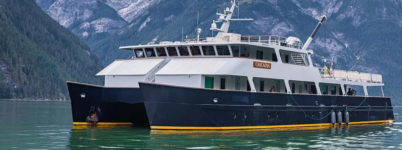 A striking expedition cruise vessel named Cascadia anchored in a pristine glacial fjord surrounded by snow-capped mountains and dense evergreen forests.