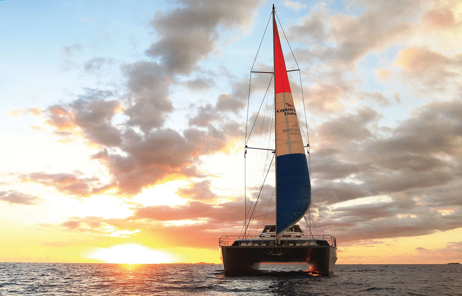 Catamaran Sailing at Tropical Sunset A modern catamaran with a red and white sail displaying 'Captain Cruse' branding sails gracefully