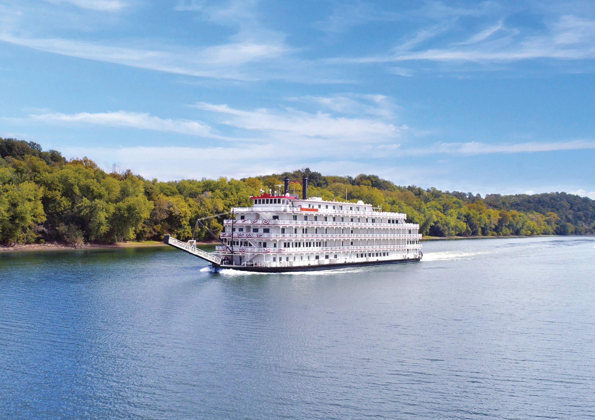 Classic Paddlewheel Riverboat on Scenic River A magnificent white multi-deck paddlewheel riverboat with red trim navigates a calm river surrounded by lush green forested banks under a bright blue sky. The vessel exemplifies classic American riverboat cruising with its traditional architecture, multiple decks, and iconic paddle wheel design.
