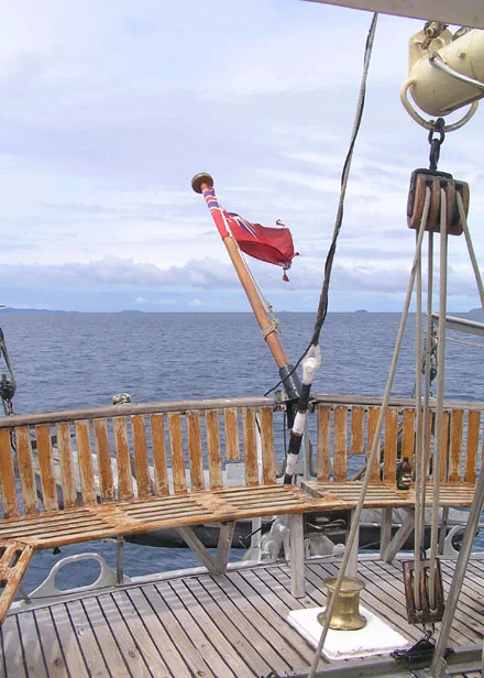 Classic Sailing Vessel on Open Ocean A traditional wooden sailing ship's deck captured from an elevated vantage point, featuring a red flag mounted on the wooden railings, maritime rigging equipment, and pulleys against a calm sea and overcast sky.