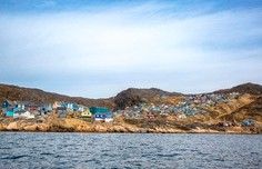 Coastal Village Harbor View A picturesque coastal settlement nestled against arid, rocky hills viewed from the water, featuring colorful buildings clustered along the shoreline with a calm sea in the foreground and blue sky above.