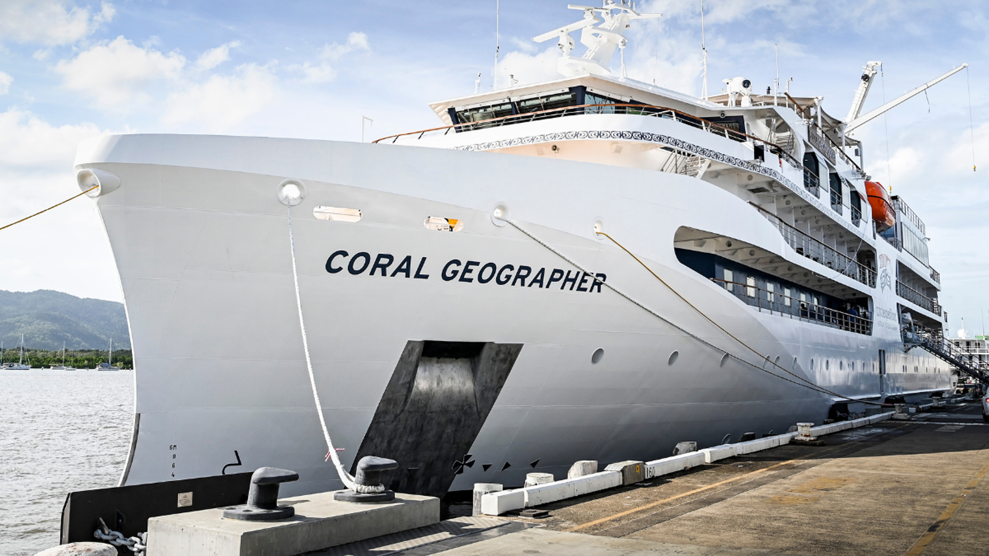 Coral Geographer Cruise Ship at Port A large white cruise ship named Coral Geographer docked at a maritime port, with mountains visible in the background and clear blue skies overhead.