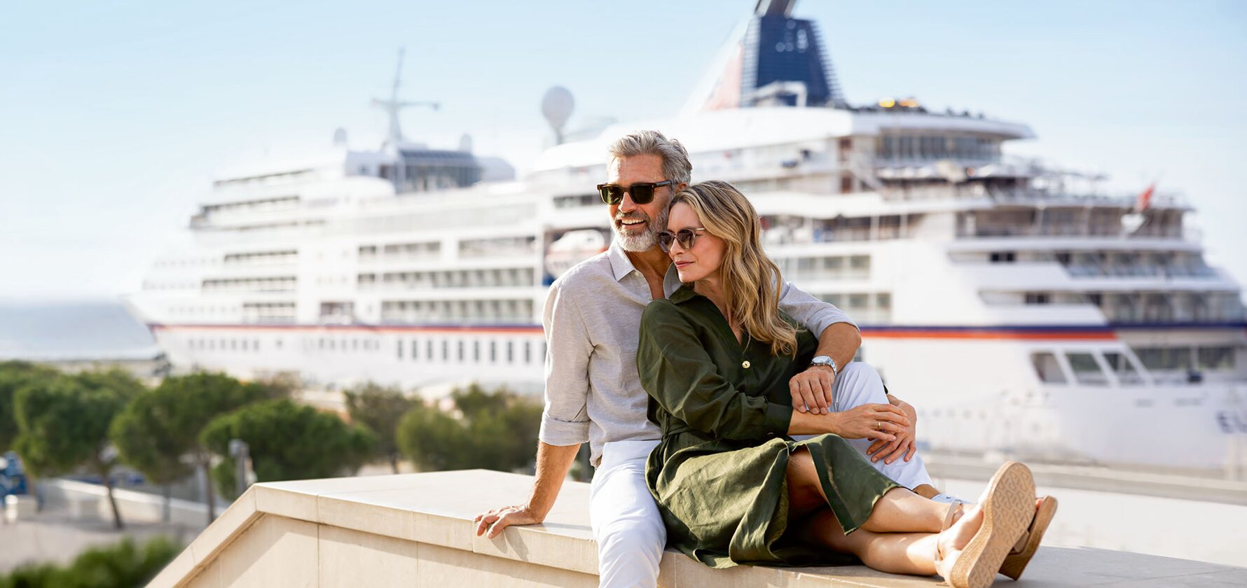 A mature couple sits together on a waterfront ledge, gazing at a large cruise ship docked in the harbor behind them.