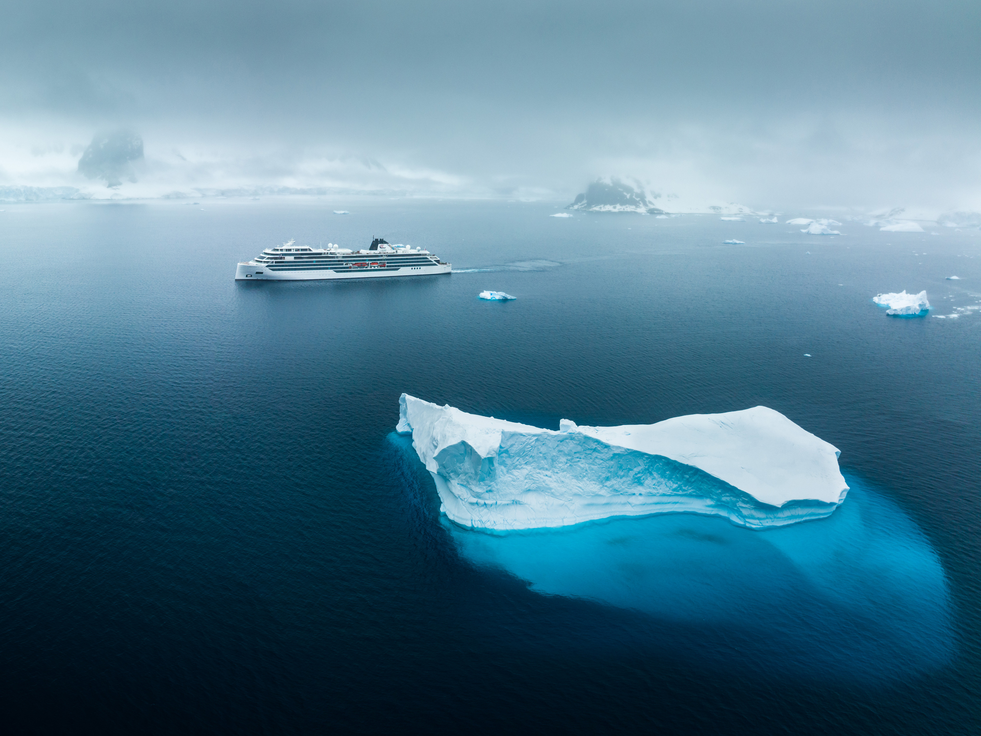 A modern white cruise ship sailing through cold arctic waters near large, blue-white icebergs with misty mountainous islands in the background.