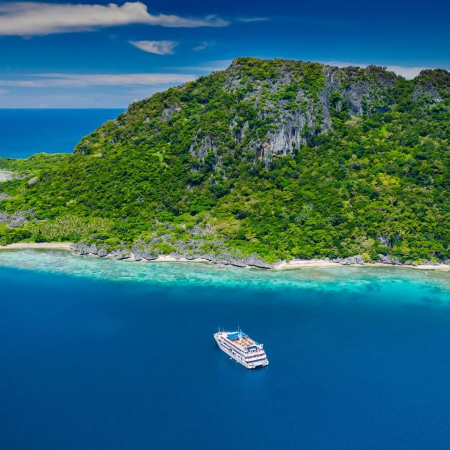 Cruise Ship Anchored at Tropical Island A modern cruise ship anchors in crystal-clear turquoise waters off a lush, forested limestone island with white sandy beaches and dramatic rocky cliffs.