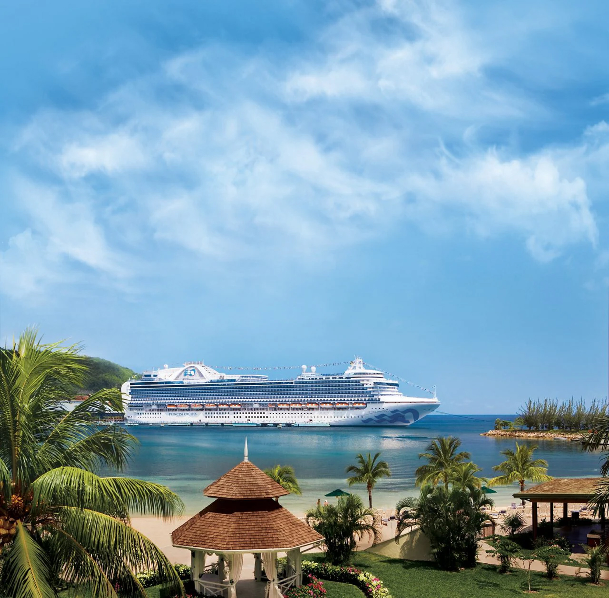 Cruise ship docked at a tropical Caribbean port with palm trees and colorful buildings in the foreground.