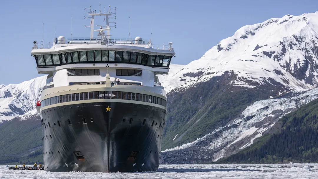 Cruise Ship in Alaskan Glacial Landscape A large cruise ship sailing through icy waters with snow-capped mountains and glacial terrain in the background. A small zodiac boat is visible near the ship's hull.