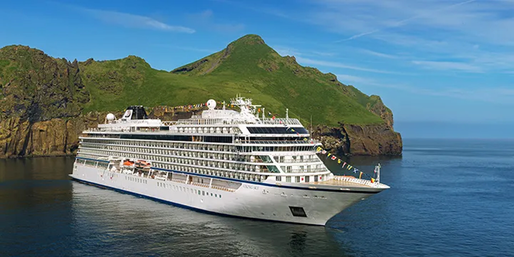 A white luxury cruise ship sailing near a green mountainous island with rocky cliffs and a calm blue ocean.