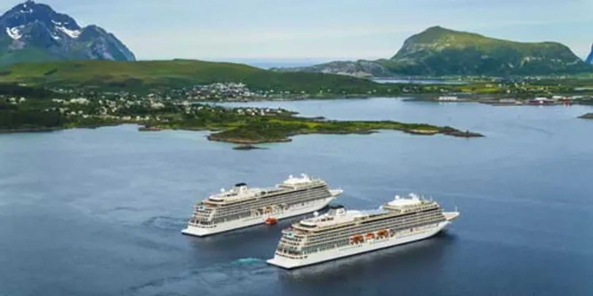 Two modern white cruise ships anchored in a picturesque Norwegian fjord, surrounded by lush green mountains and a small coastal town in the background.
