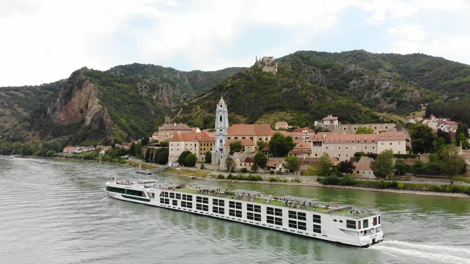 Danube River Cruise in Historic European Landscape A modern white river cruise ship sailing along the Danube River, passing a picturesque historic town nestled against rocky forested hills with a church tower and ancient castle in the background.