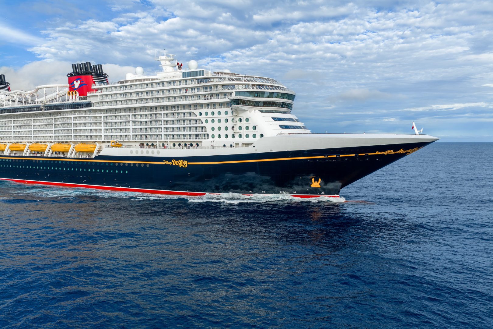Disney Cruise Line Cruise Ship at Sea A massive modern cruise ship with distinctive Disney branding on its red funnel featuring Mickey Mouse, anchored in calm blue waters under a partly cloudy sky.