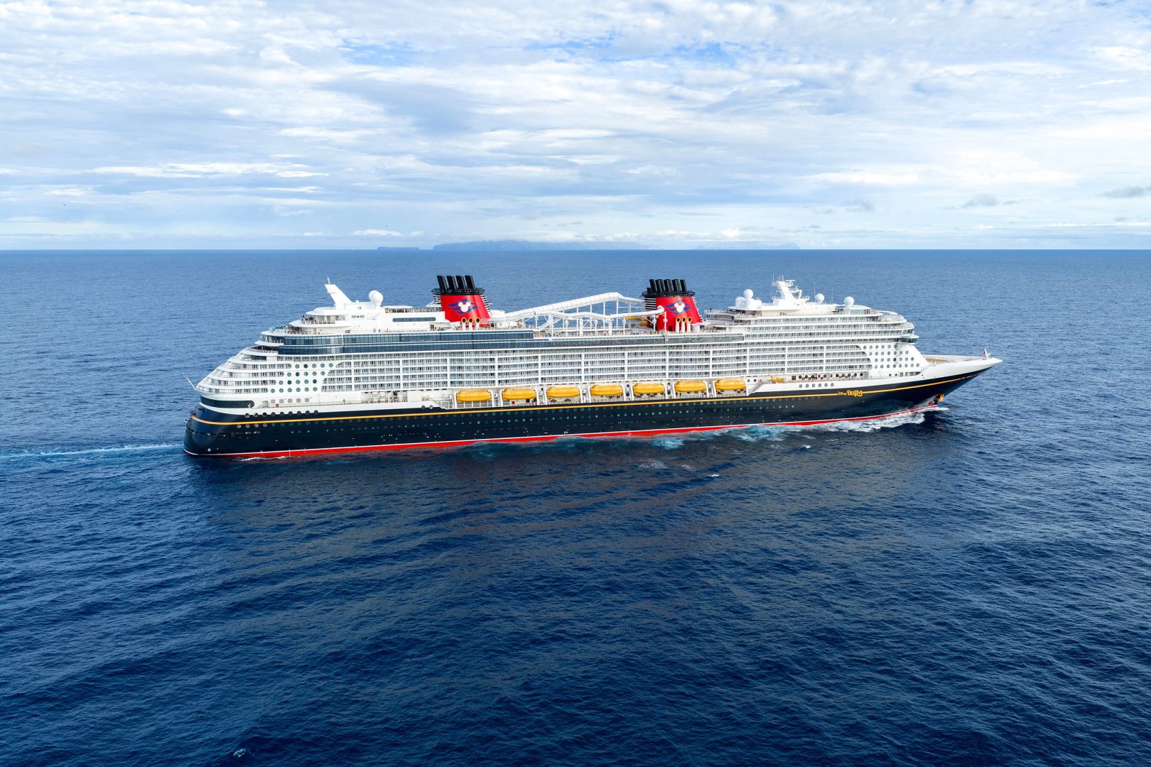 Disney Cruise Line Ship at Sea A massive contemporary cruise ship navigates open ocean waters under a partly cloudy sky with distinctive red funnels and dark blue hull.