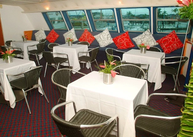 Elegant Dining Room with River Views A sophisticated cruise ship dining venue featuring round tables with white linens, woven rattan chairs, and red decorative pillows, overlooking a scenic waterway through expansive windows.