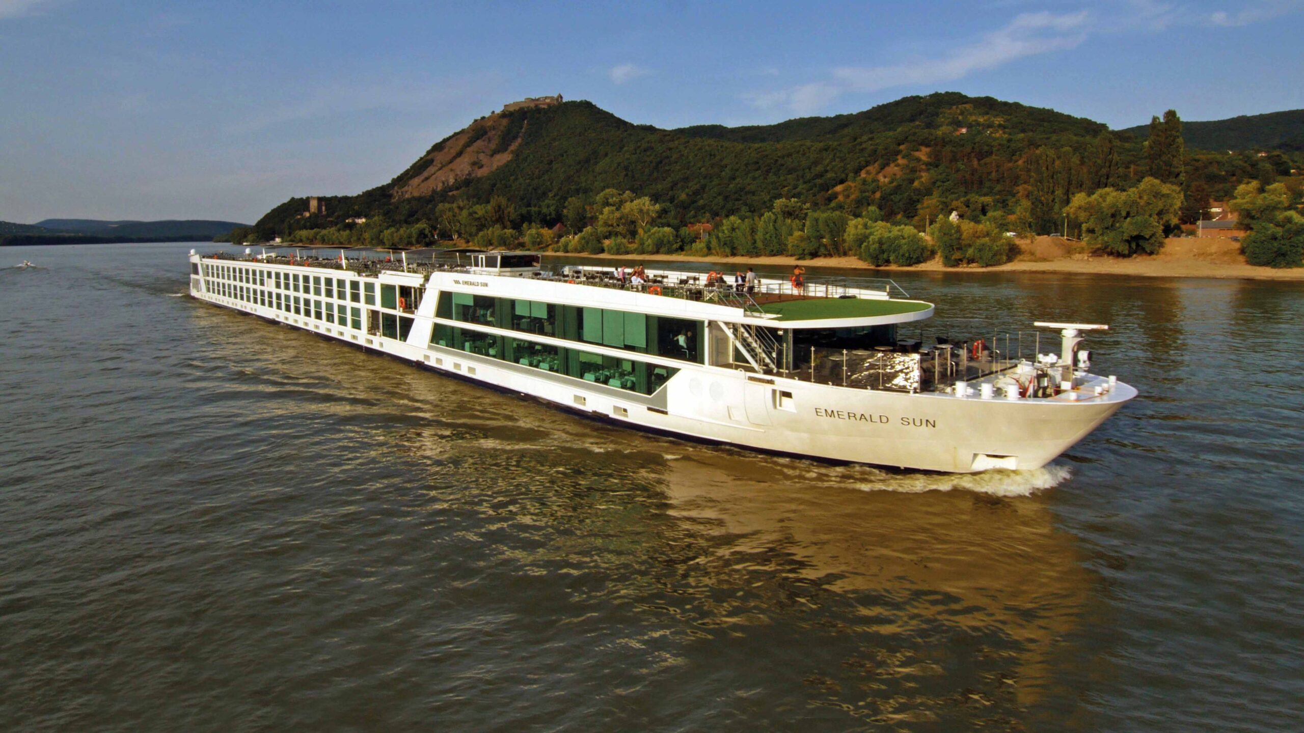 A river cruise ship navigates through a lush, forested river valley with steep hillsides, passing picturesque towns and winding roads along the riverbanks.