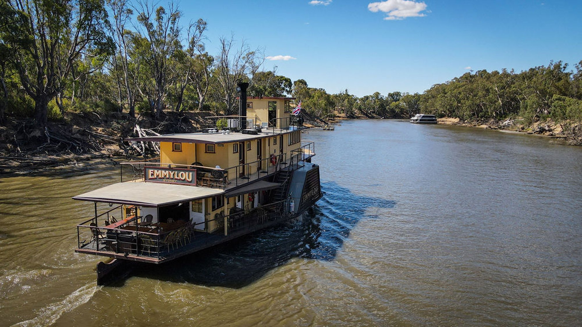 A charming paddle-wheeler riverboat named Emmylou moored along the Murray River in Australia, featuring a distinctive yellow and cream-colored two-story design with open deck areas. The vessel is anchored in shallow water surrounded by native eucalyptus trees and arid landscape under clear blue skies.