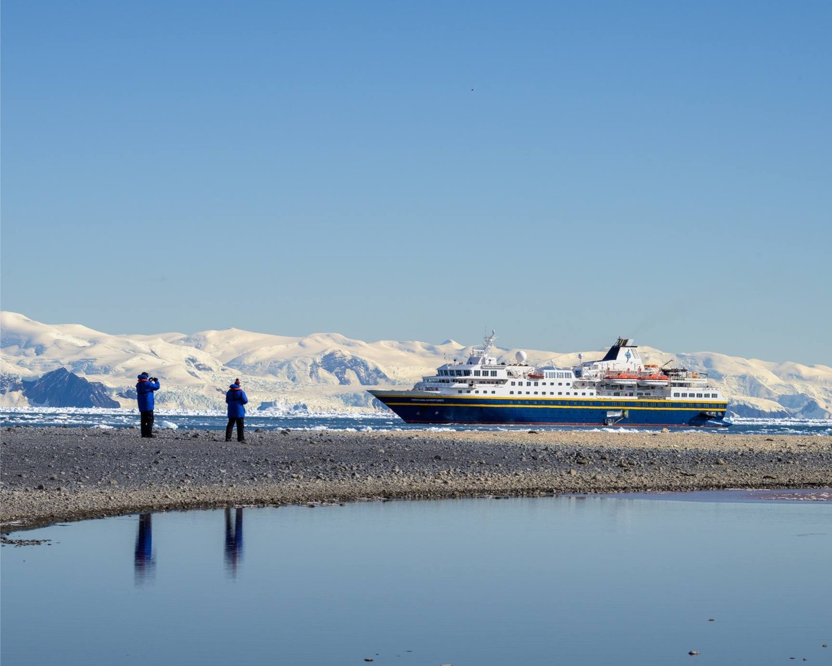 Expedition Cruise Ship in Antarctic Waters A large expedition cruise ship is anchored in pristine Antarctic waters, with passengers on the rocky beach observing the vessel and snow-covered mountains in the background.