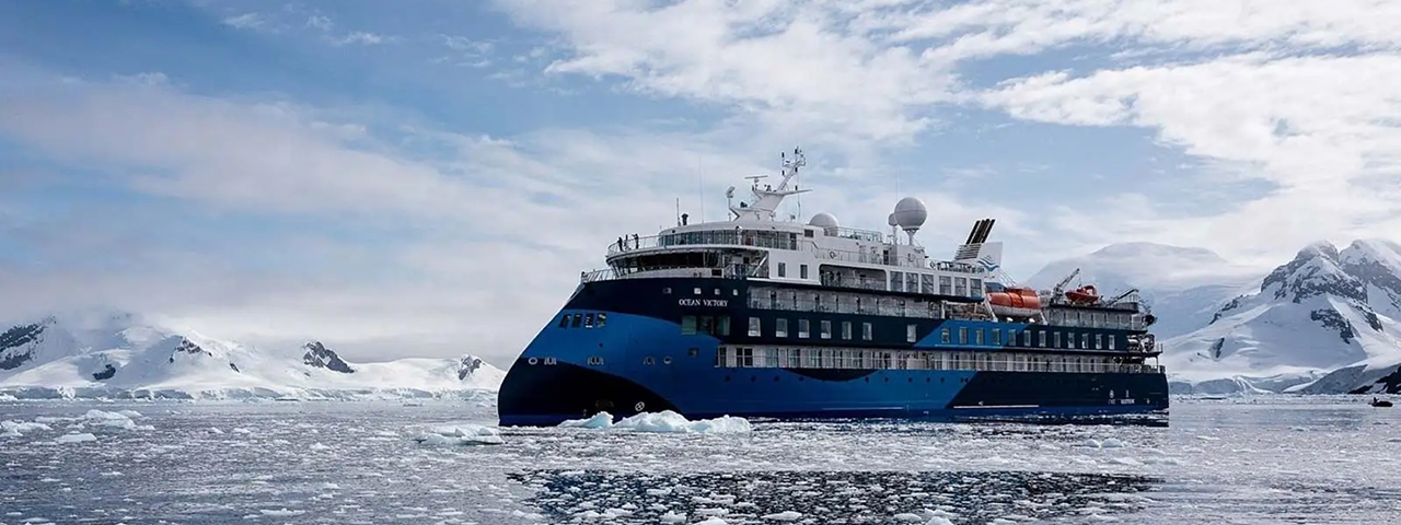 A modern expedition cruise ship with distinctive blue and black hull navigates through icy Antarctic waters surrounded by snow-capped mountains and floating ice.