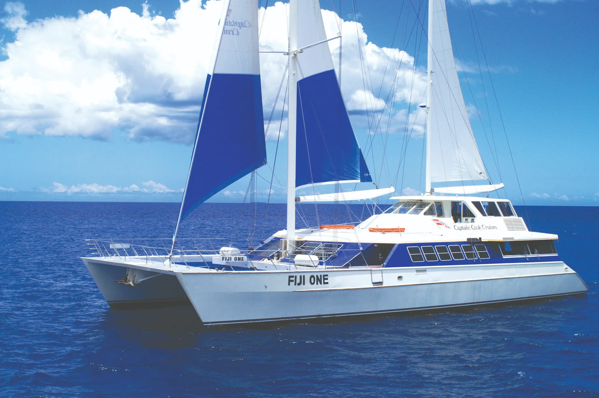Fiji One Sailing Catamaran Cruise The Fiji One, a modern sailing catamaran operated by Captain Cook Cruises, glides across crystalline blue waters with billowing white and navy sails deployed.