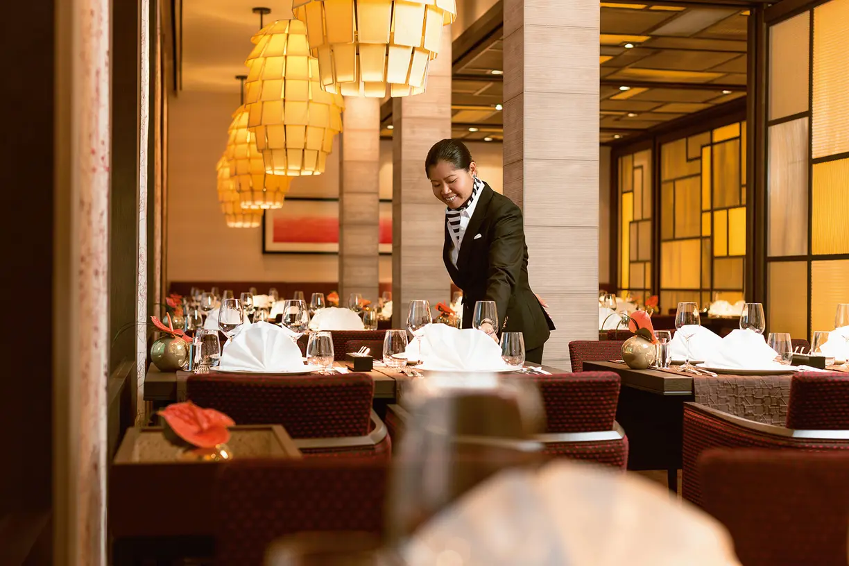 A professional server prepares an elegant dining table in a high-end restaurant with warm golden lighting, white linens, and sophisticated place settings.