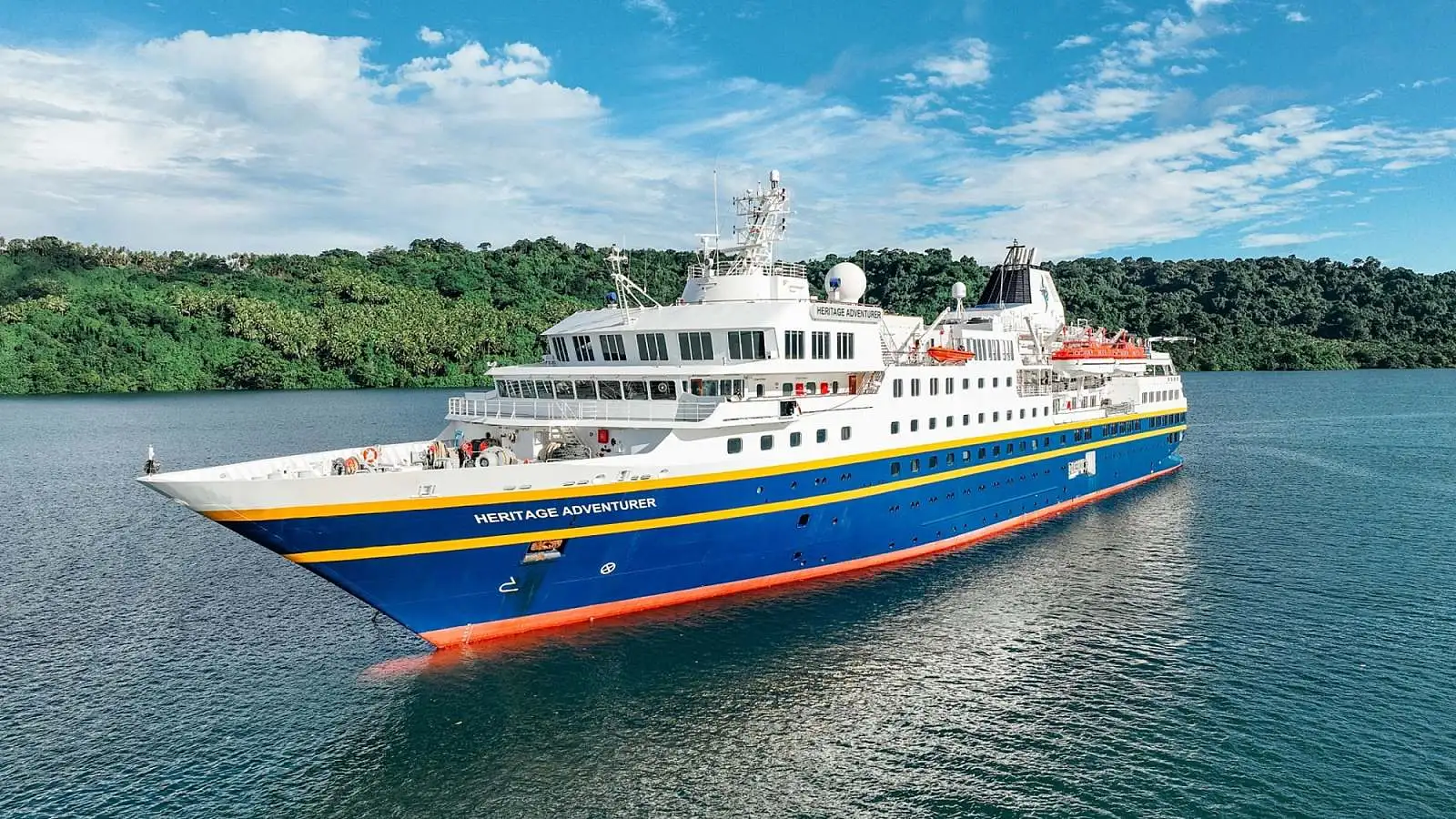 The Heritage Adventurer, a modern expedition cruise ship with distinctive blue and orange hull livery, anchors in turquoise tropical waters surrounded by lush green mangrove forests.