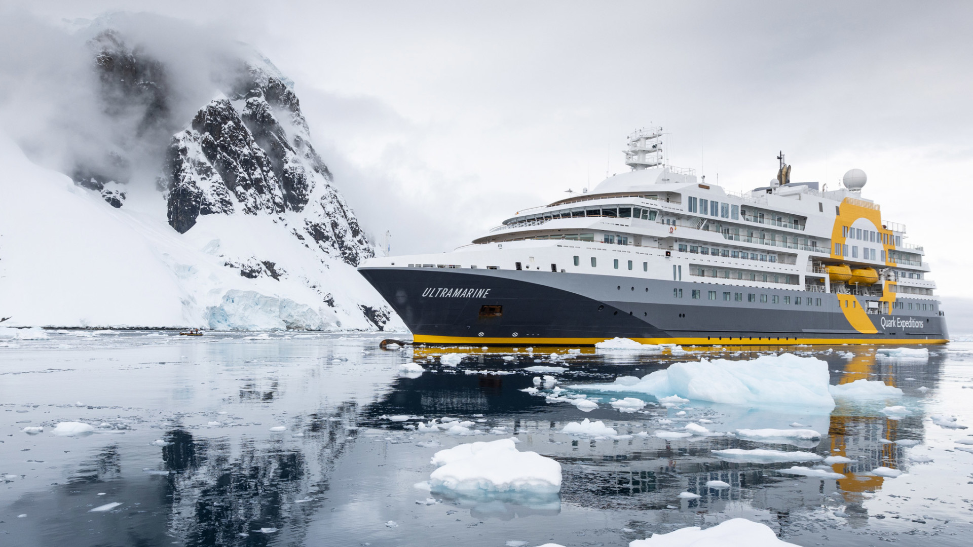 Hero_Sam-Crimmin-V3-3702 The Ultramarine expedition cruise ship navigating through icy Antarctic waters surrounded by snow-capped mountains and floating icebergs.