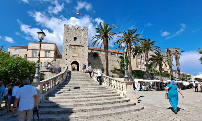 Historic Stone Tower in Mediterranean Coastal Town A scenic plaza featuring a medieval stone tower with a flag at its peak, connected by a grand