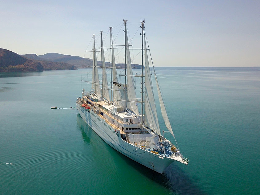 Luxurious Sailing Yacht in Calm Turquoise Waters A stunning aerial view of a large multi-masted sailing cruise ship anchored in pristine turquoise waters near a mountainous coastal region. The vessel features multiple tall masts and white sails, with mountains visible in the background across calm, crystalline seas.