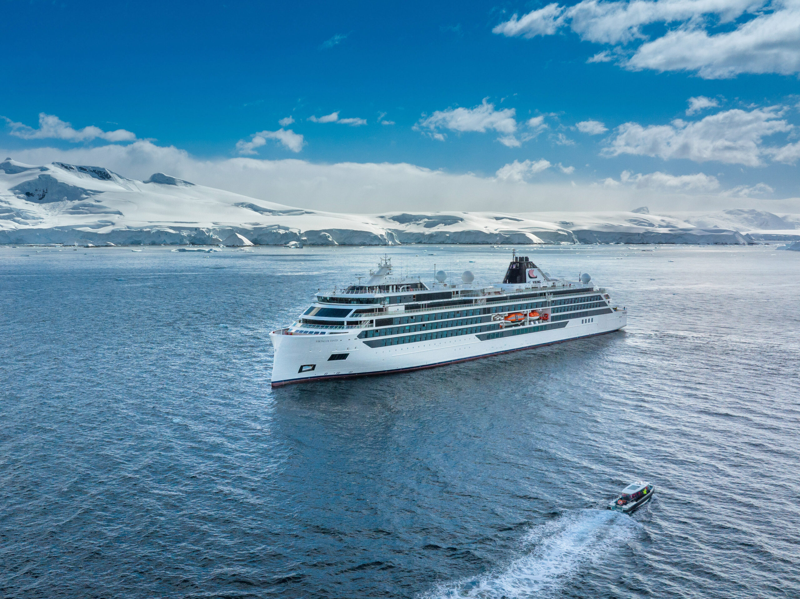 A pristine white luxury cruise ship navigating through icy blue waters with snow-covered mountainous Antarctic landscape in the background.