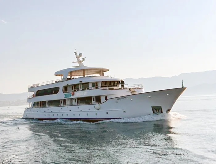 Luxury Motor Yacht Anchored in Tropical Waters A stunning white multi-deck luxury yacht anchored in crystal-clear turquoise Mediterranean waters under a bright blue sky. The vessel features elegant modern architecture with multiple passenger decks and appears to be a premium cruise or charter yacht positioned in a pristine tropical or subtropical coastal location.