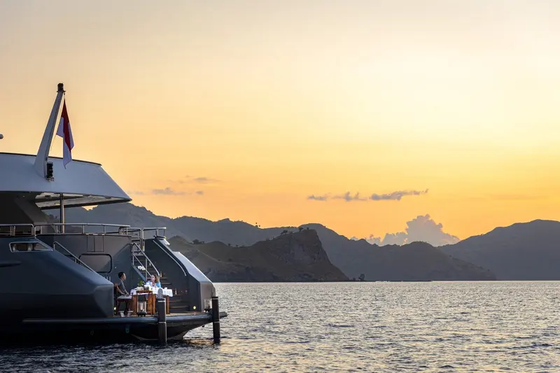 Luxury Yacht at Tropical Sunset A modern luxury yacht tender is moored at sunset with dramatic golden-orange skies reflecting off calm waters, with mountainous tropical islands silhouetted in the background.