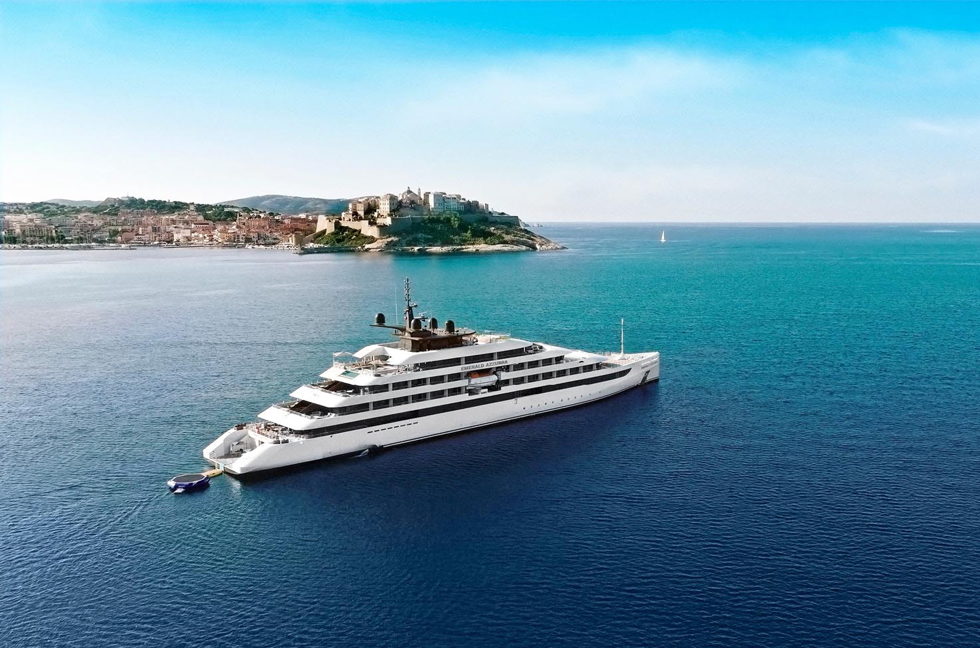 A sleek, modern white luxury yacht anchored near a historic coastal town with stone buildings perched on a green peninsula, surrounded by turquoise Mediterranean waters.