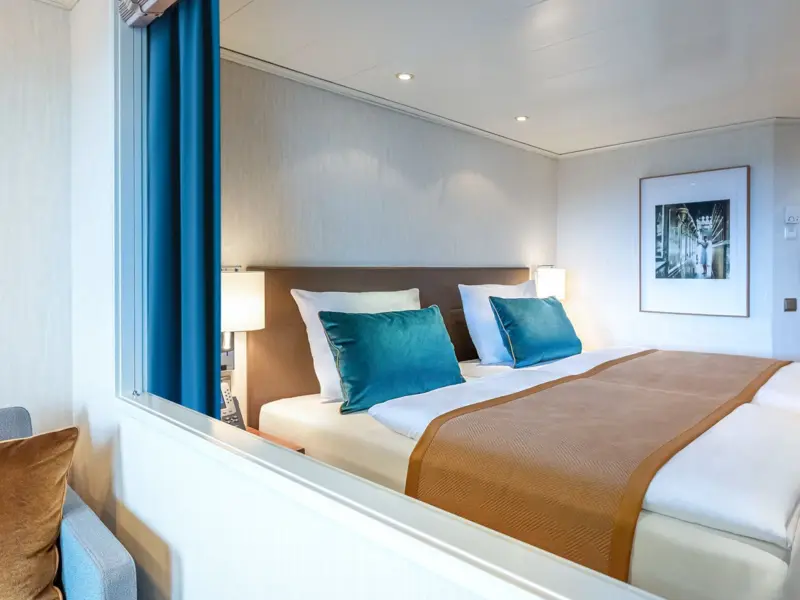 A contemporary cruise ship stateroom featuring a comfortable double bed with teal accent pillows, warm wood furnishings, and a large picture window with ocean views.