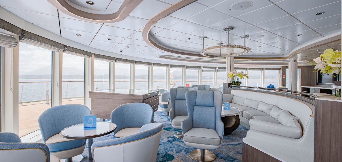 A sophisticated and contemporary lounge area aboard a luxury cruise ship featuring floor-to-ceiling windows with panoramic ocean views, stylish blue and gray upholstered seating, modern pendant lighting, and elegant architectural details including curved ceiling elements and wooden accents throughout the space.