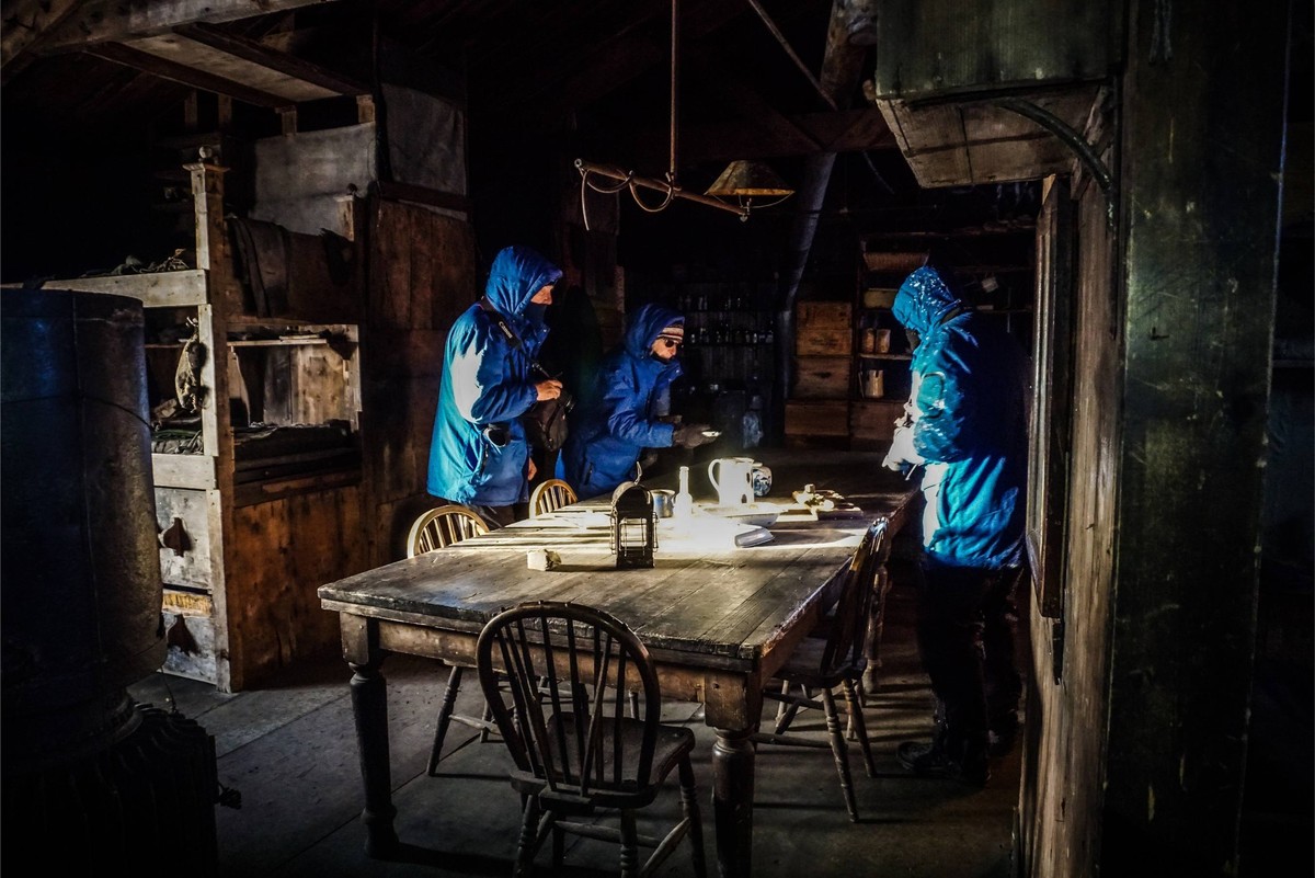 Night Gathering in Historic Industrial Space Three people in bright blue jackets gather around a weathered wooden table in a dimly-lit, rustic