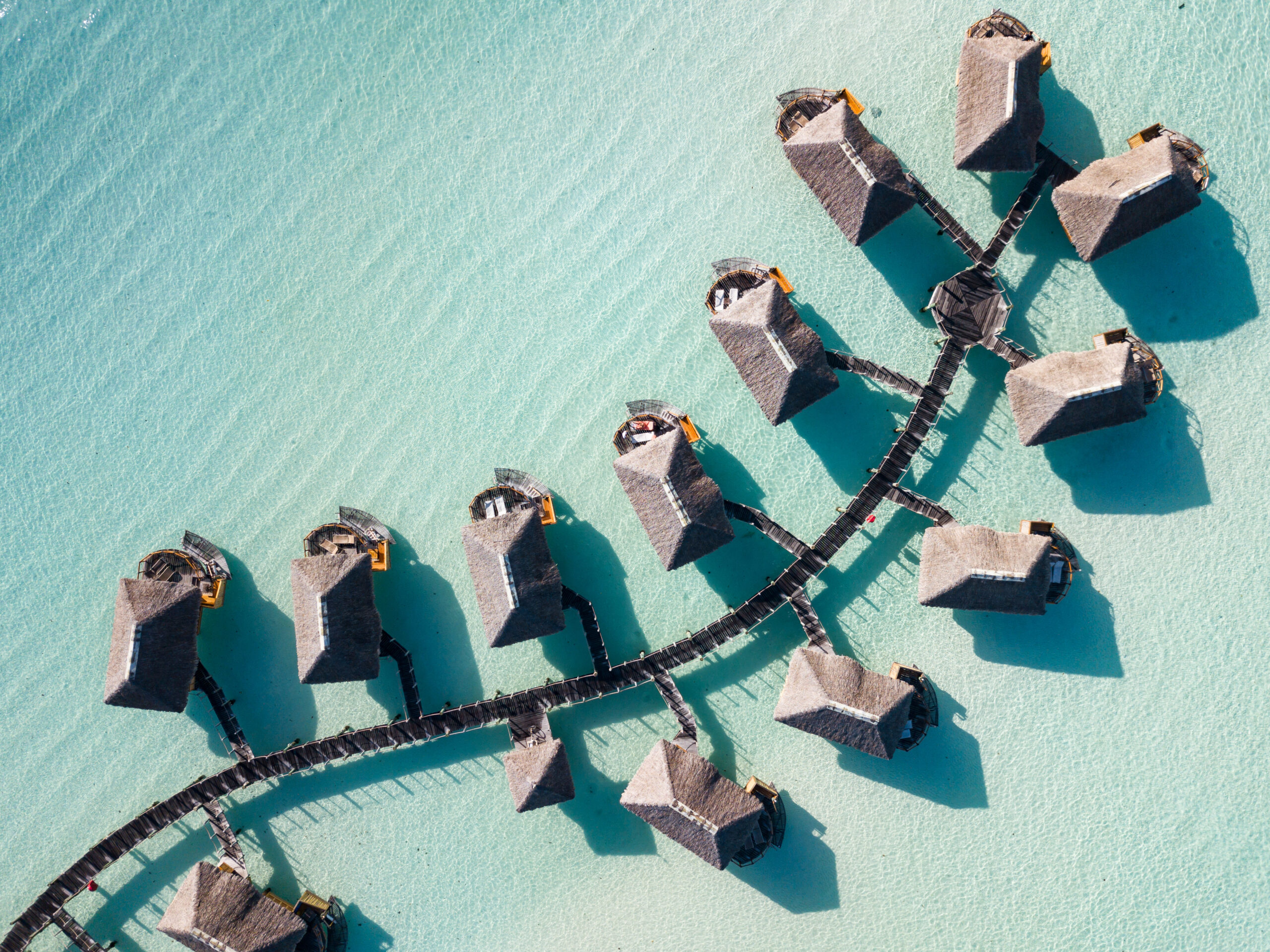 Overwater Bungalows Aerial View An aerial view of a luxury resort with thatched-roof overwater bungalows arranged in a curved pattern along wooden walkways in crystal-clear turquoise lagoon waters.