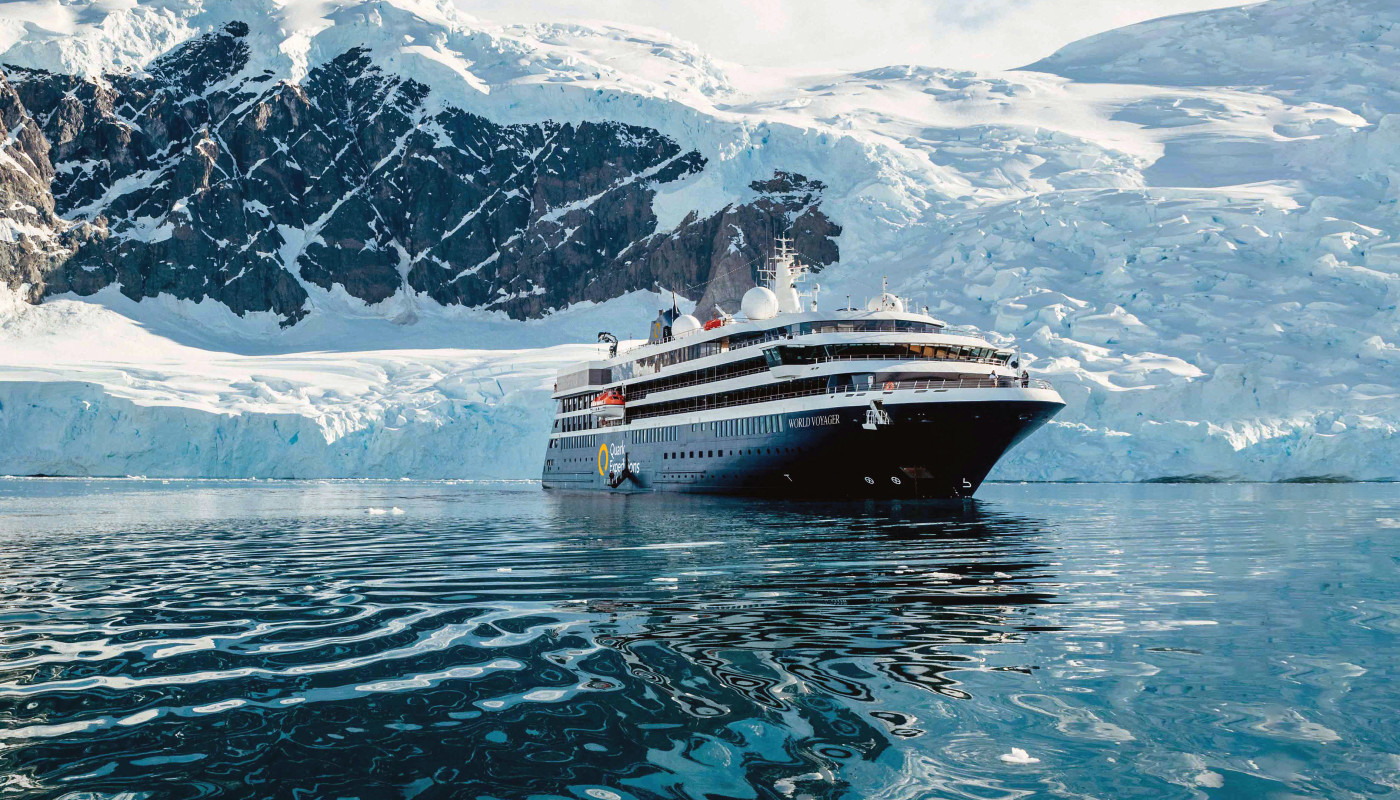 QE_WVO_Exterior-Shots_1 A modern expedition cruise ship navigating through icy waters with snow-covered terrain and a helicopter overhead, showcasing polar exploration capabilities.