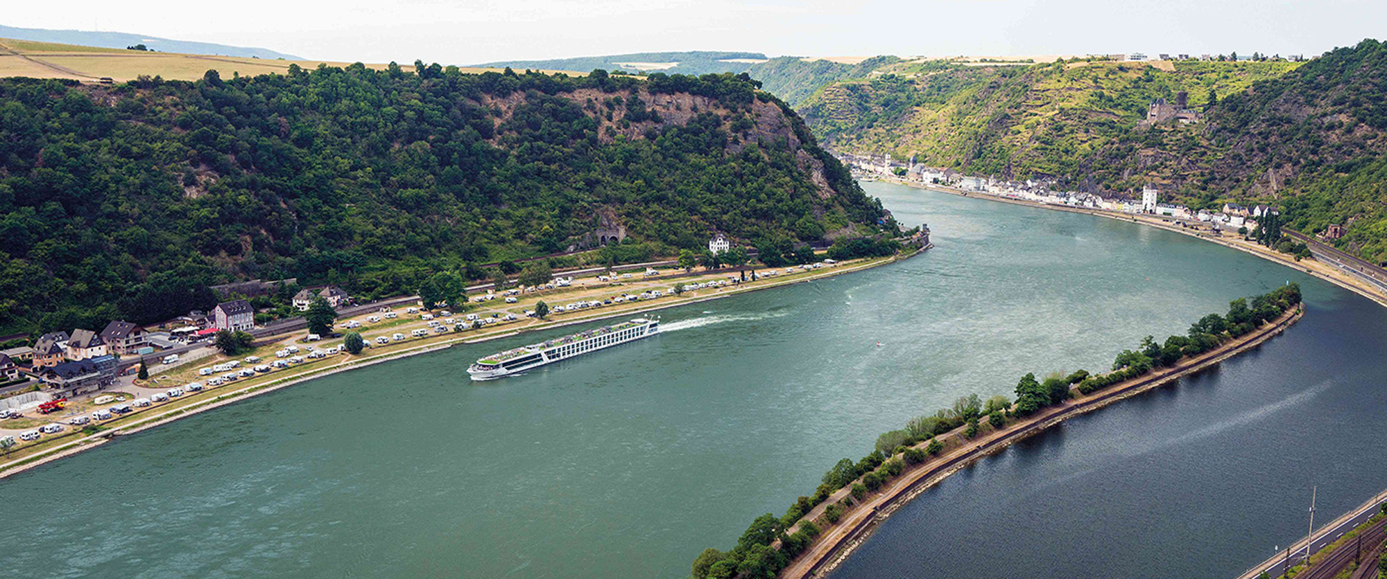 A scenic aerial view of a river cruise ship navigating through the dramatic Rhine Gorge, with steep forested cliffs, a charming riverside village, and lush green hillsides.