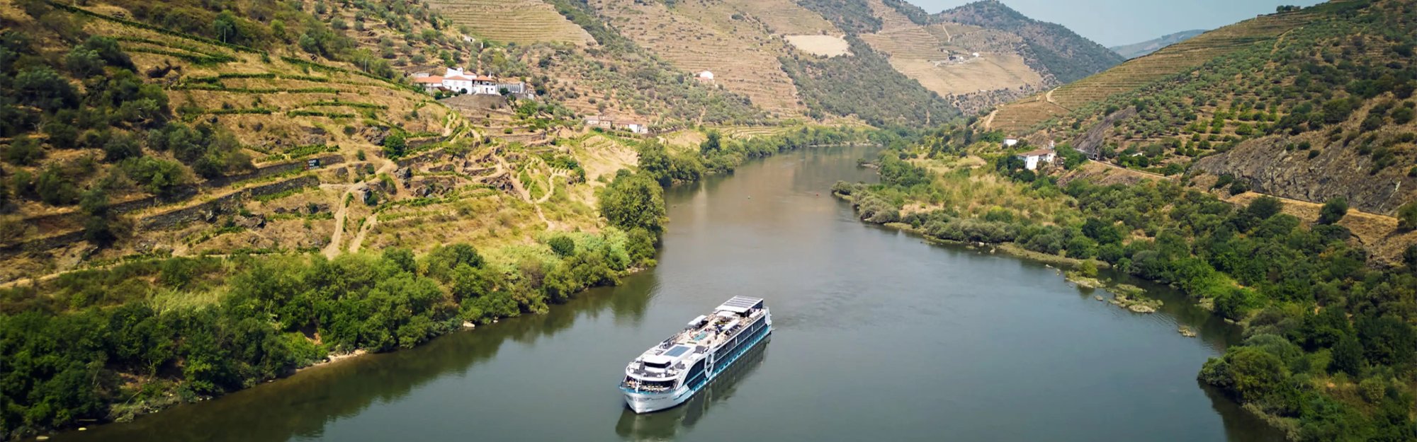 River_Cruise_Hero_edit A modern river cruise ship navigates through the scenic Douro River valley, surrounded by terraced vineyards on steep hillsides and traditional villages perched on the landscape.
