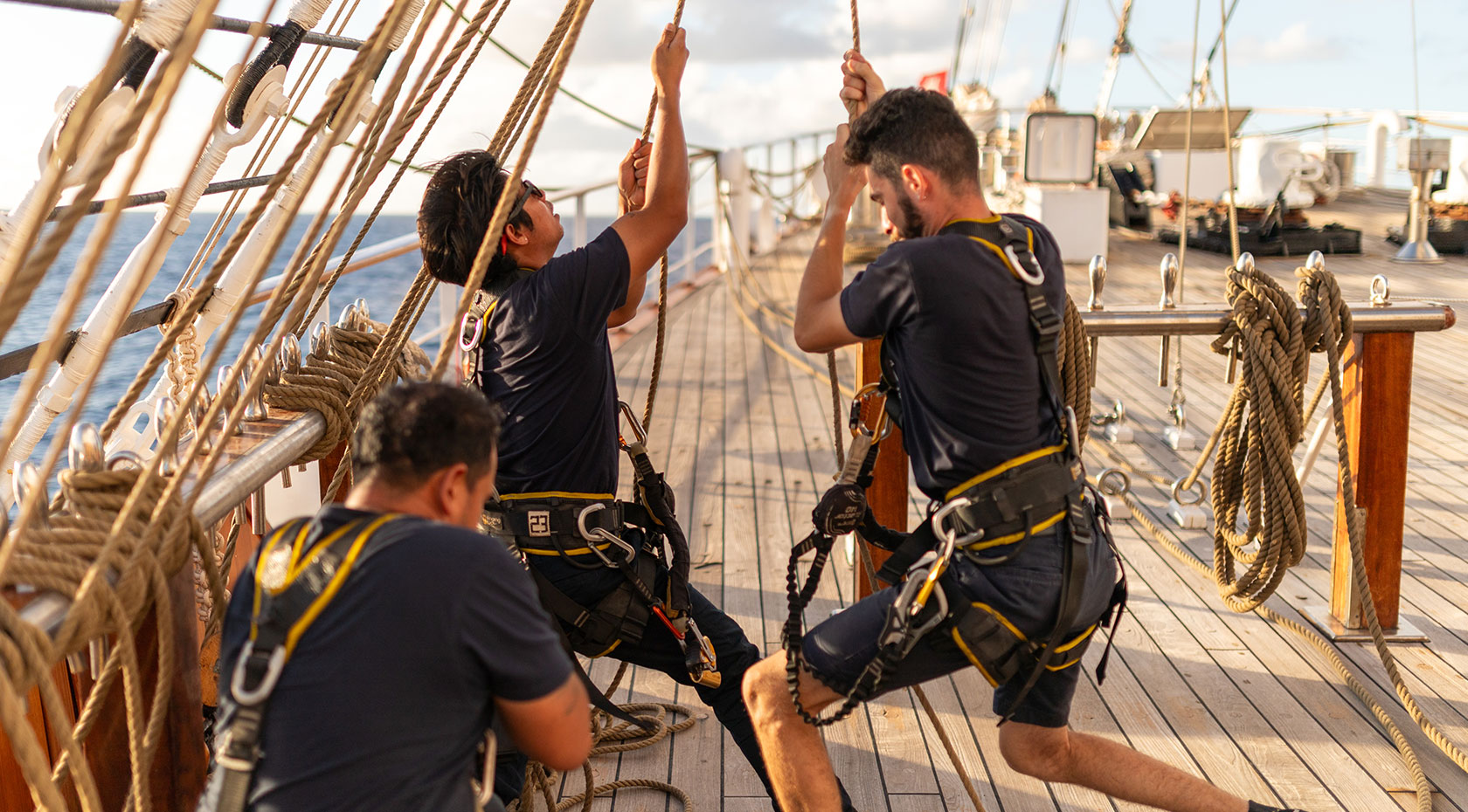 SCII_PE_Crew_002_1680x930 A team of crew members in dark uniforms with yellow accents work together on the deck of a traditional tall ship, adjusting ropes and rigging.