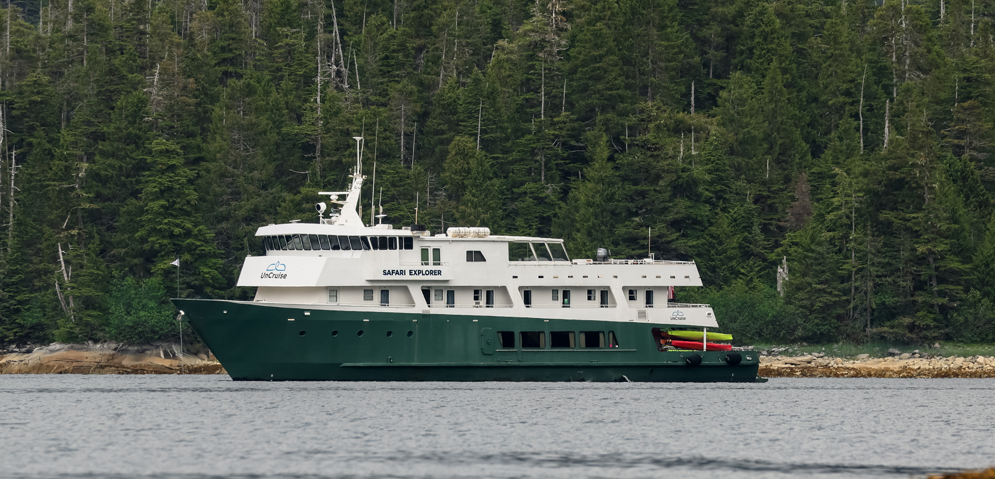 Safari Explorer Expedition Cruise Ship A green and white expedition cruise ship named Safari Explorer anchored near a dense evergreen forest coastline, with colorful kayaks visible on its side.