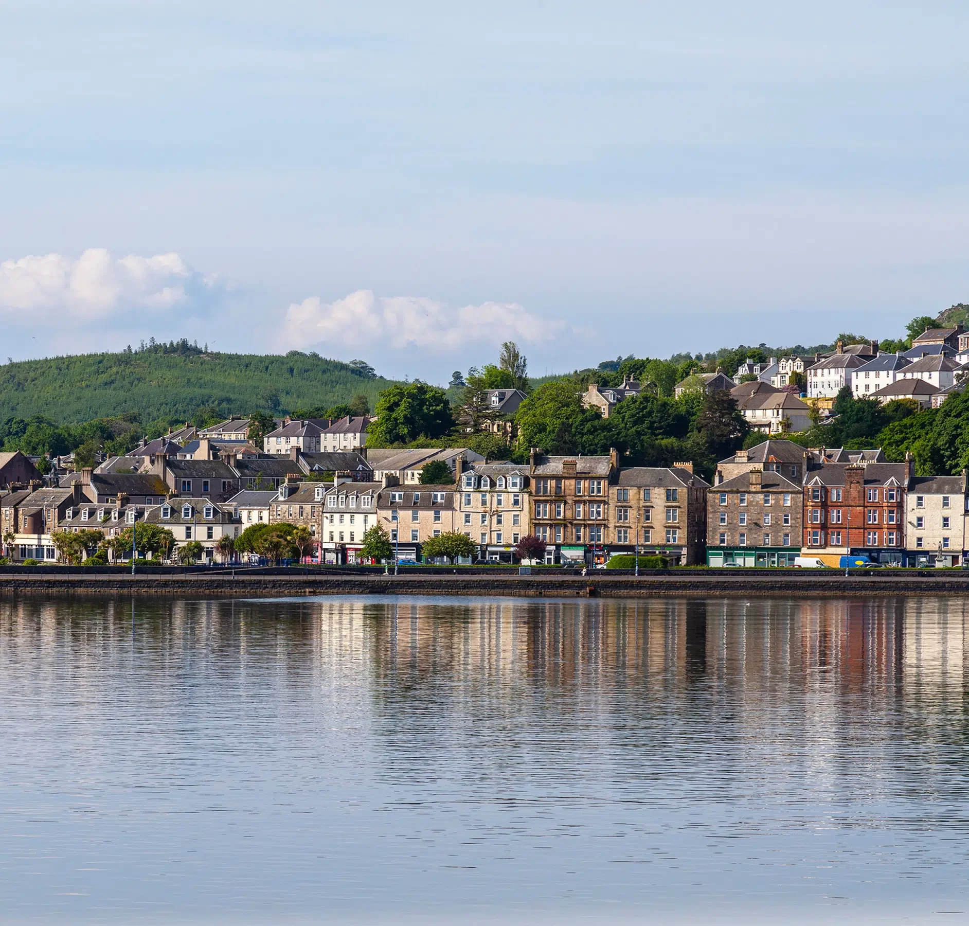 Scottish Riverside Town Waterfront View A picturesque Scottish riverside town featuring a row of traditional Georgian and Victorian stone buildings reflected in calm waters, with forested hills rising in the background.