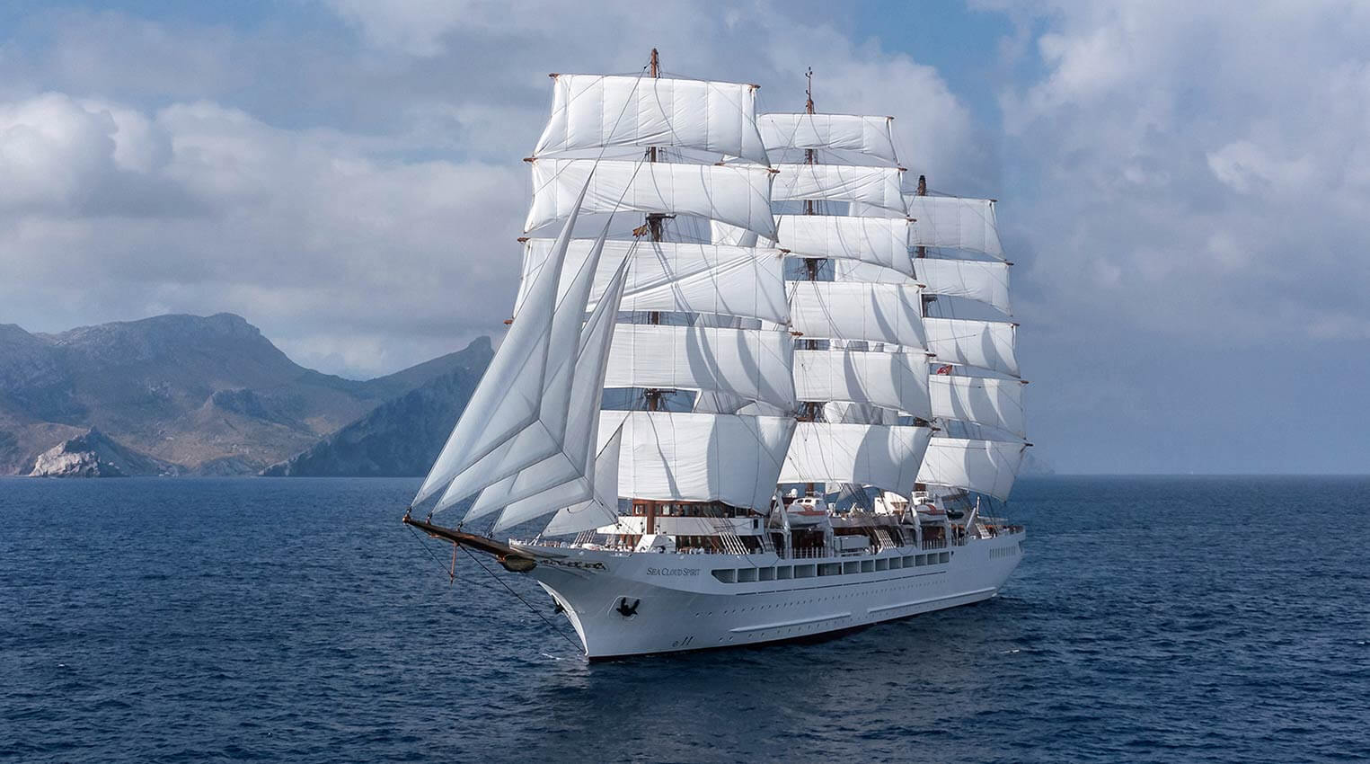 SeaCloud_Kreuzfahrten_SeaCloudSpirit_Schiff4 A magnificent full-rigged tall ship with white canvas sails deployed, sailing in blue Mediterranean waters with a mountainous coastline visible in the background.