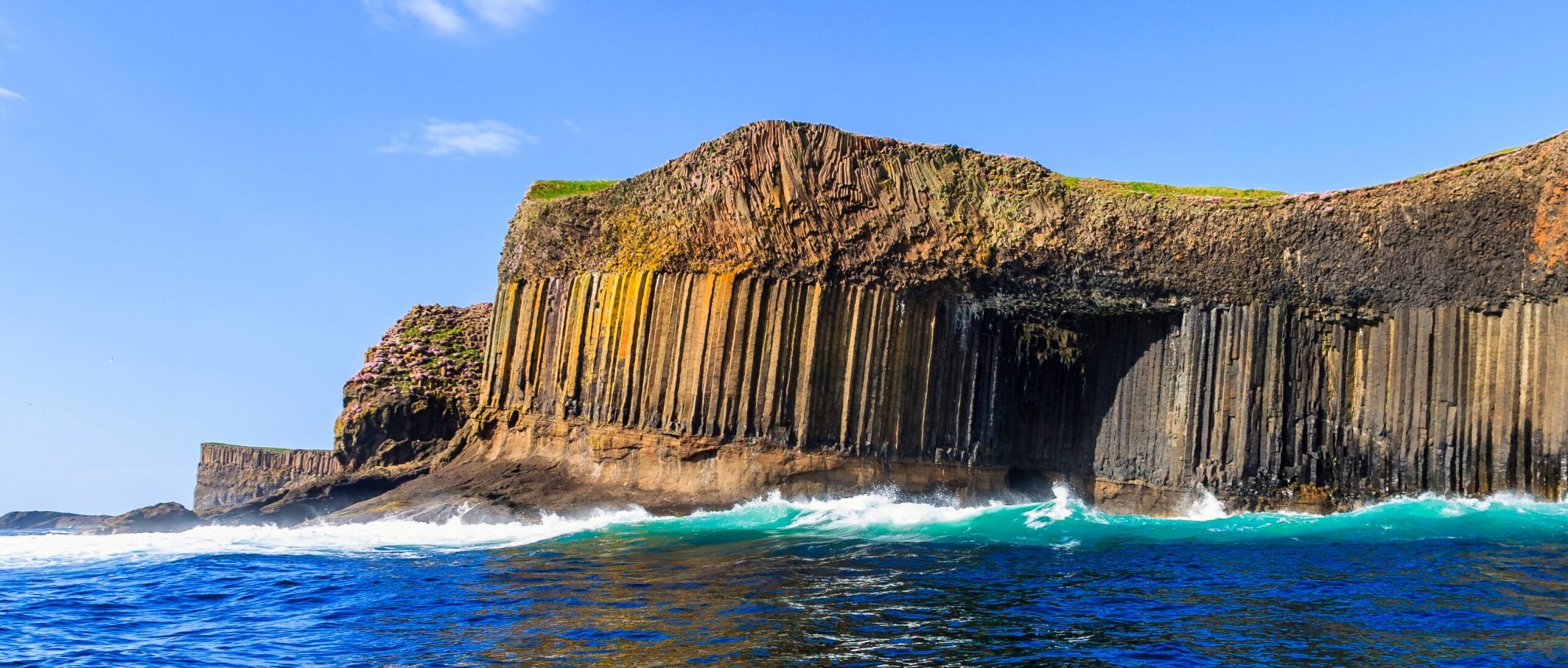 Staffa Basalt Columns and Coastal Cliffs A dramatic coastal landscape featuring distinctive hexagonal basalt columns rising from turquoise waters, with waves crashing against the volcanic rock formations. The scene showcases the UNESCO-recognized geological formations of Staffa in Scotland.